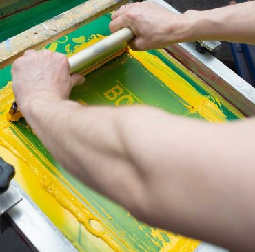 Serigraphy silk screen print process at clothes factory. Frame, squeegee and plastisol color paints.